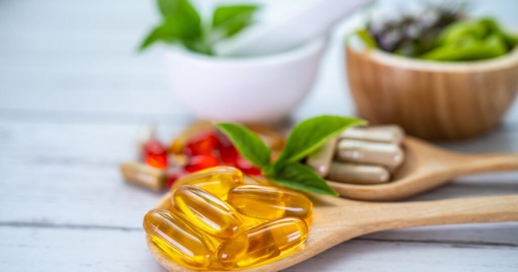 Soft gel vitamin capsules resting on two wooden spoons with fresh basil leaves and blurred bowls in the background